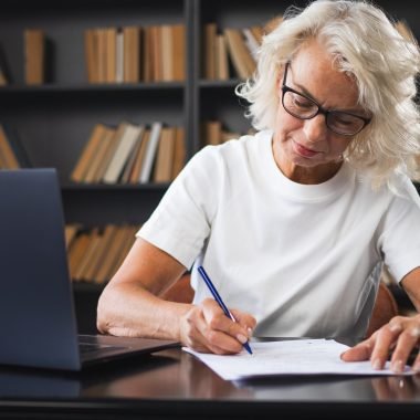 Middle aged senior woman using laptop computer writing notes at workplace. Focused mature old businesswoman work at office making written records doing online work. Mature boss professional worker
